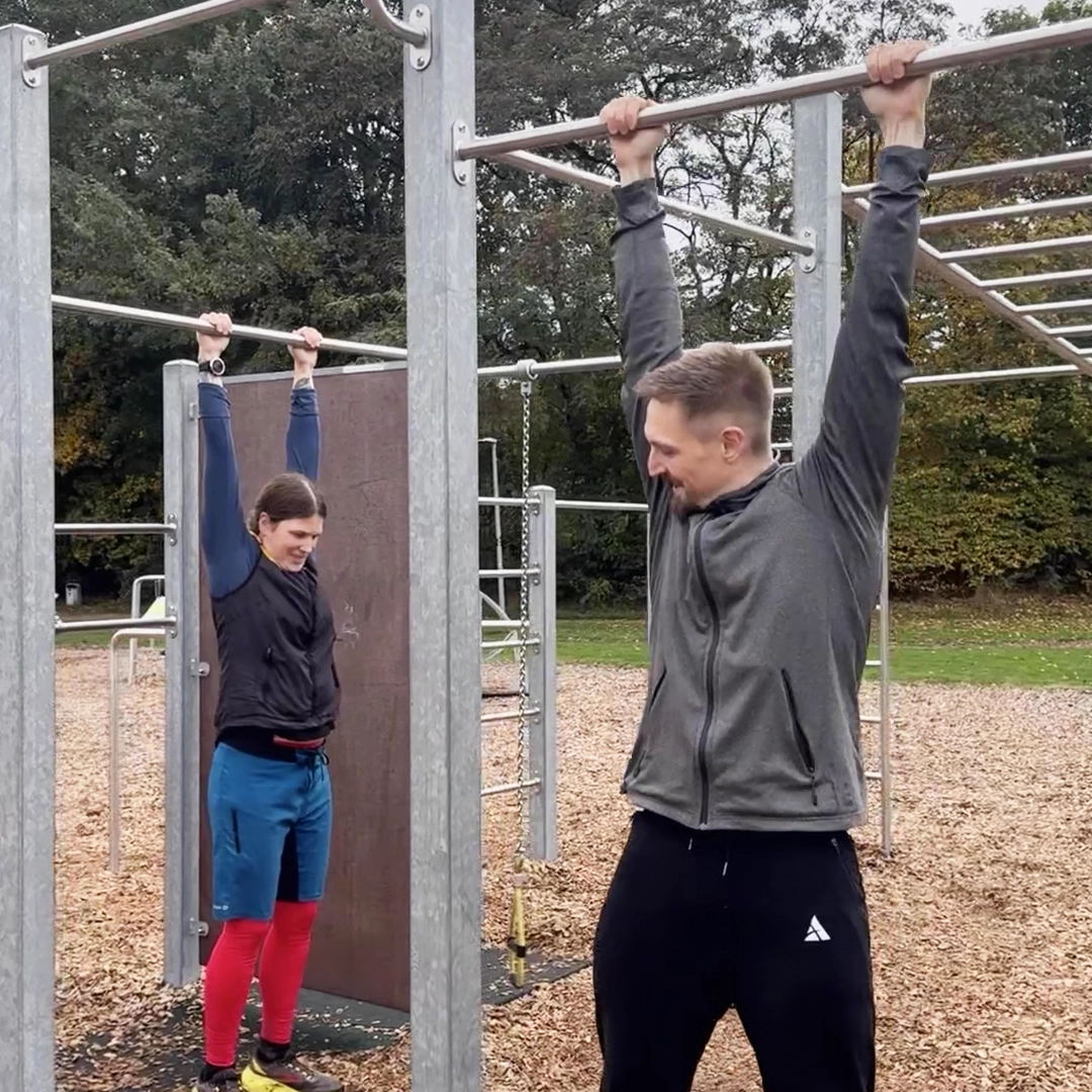 Ein Mann und eine Frau hängen an einer Stange in einem Calisthenicspark.