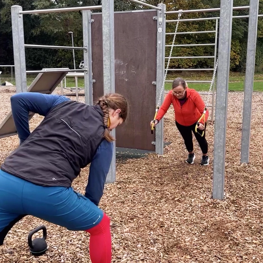 Zwei Frauen sind in einem Calisthenicspark. Eine macht einen Row mit der Kettlebell und die andere macht Push Ups an Schlingentrainern.